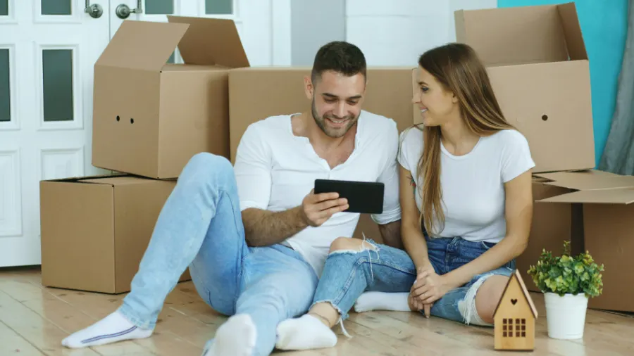 A young couple sitting on the floor among moving boxes, looking at a tablet together